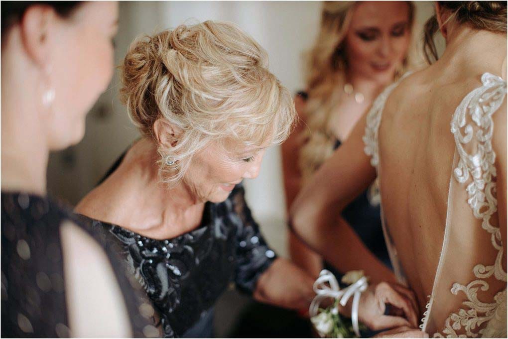 Mother of the bride helps the bride get into her dress, zipping it up in the back.