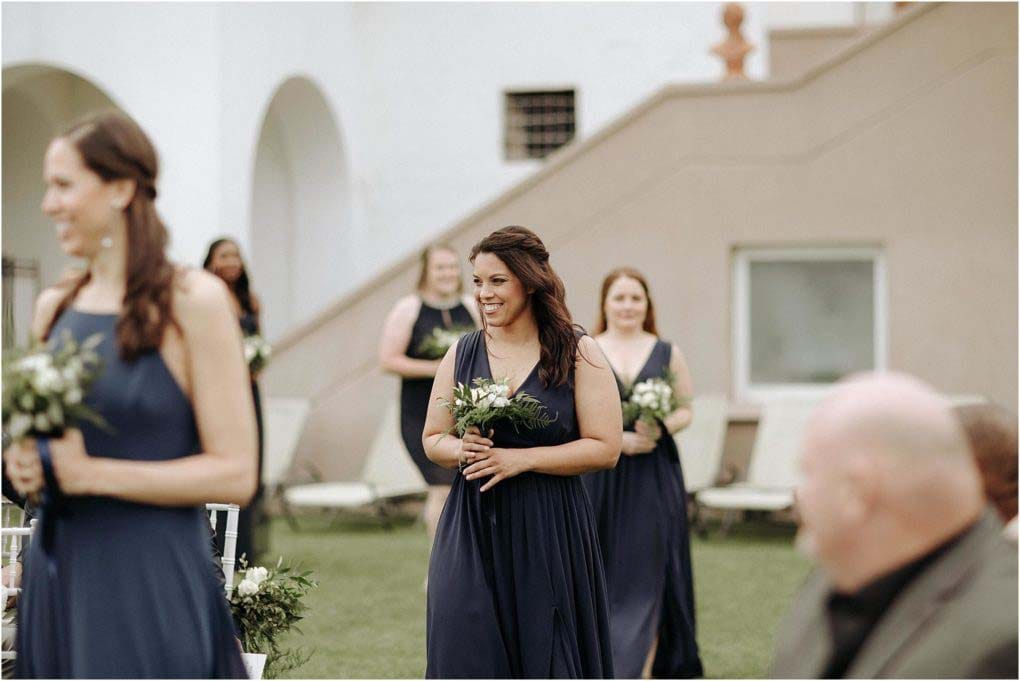 Bridesmaids enter the ceremony holding small bouquets and wearing navy blue dresses.