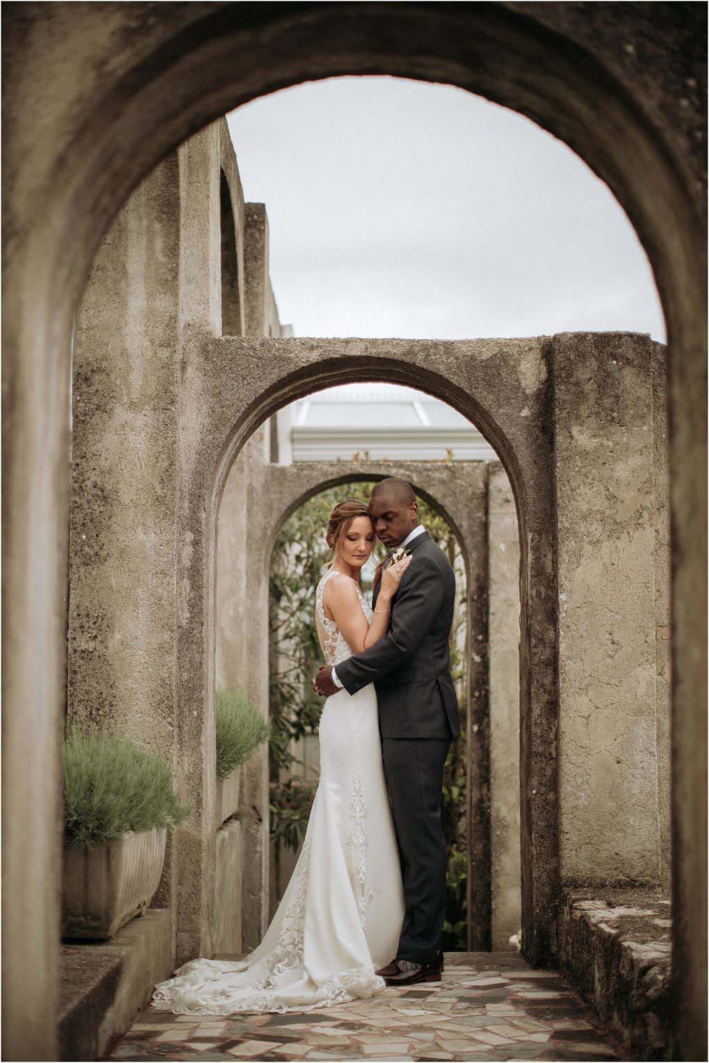 Bride and groom stand under an Italian archway, embracing.