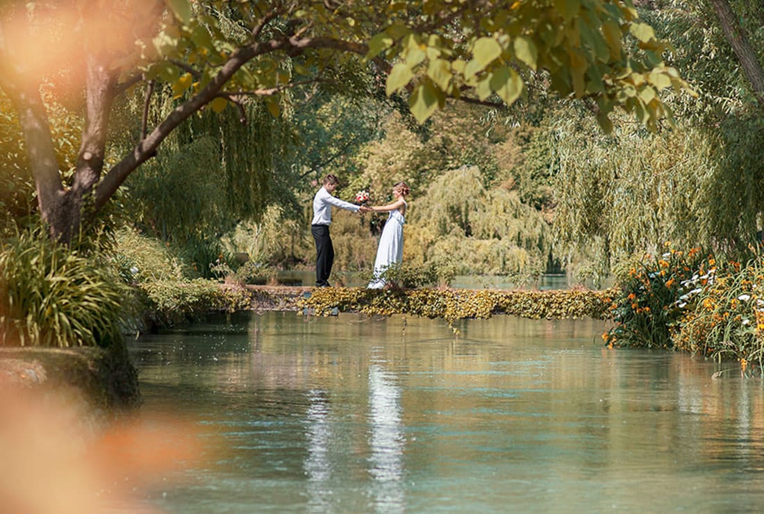 Bride and groom stand by the lake holding hands.