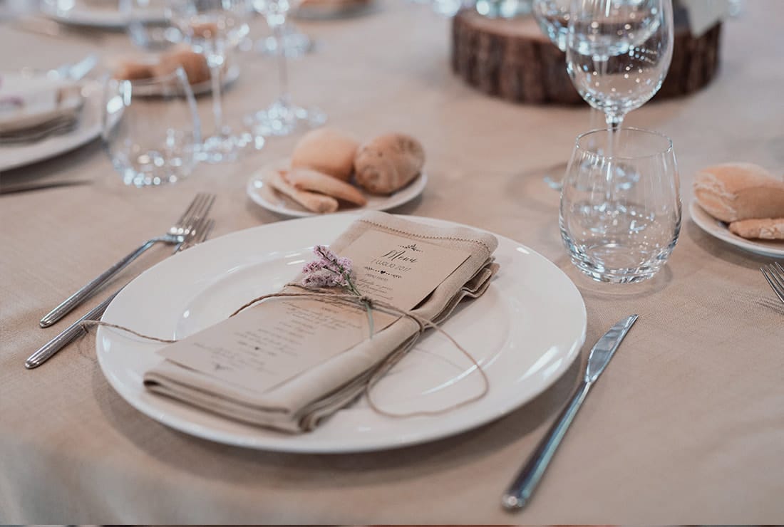 A place setting for a wedding reception. There is a water glass, a wine glass, a white plate with a napkin fold, silverware and a bread plate at the top left corner.