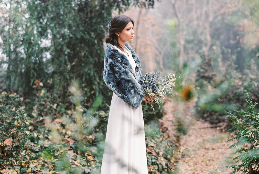 A bride standing in a wooded area holding her blue thistle bouquet and wrapped in a blue fur. She is wearing a white wedding dress and has dark hair.