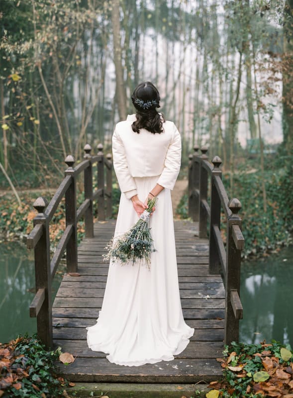 A winter bride with a fur wrap stands on the edge of a bridge over the pond. She holds her bouquet behind her back. 