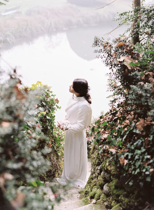 A winter bride stands on a small path overlooking a pond. She is surrounded with vines and greenery.