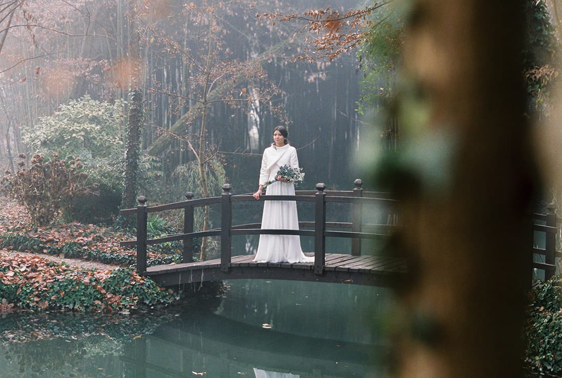 A winter bride stands on a bridge over a small pond. She is standing beneath bare trees and holding her bridal bouquet. She wears a white gown with a fur wrap.