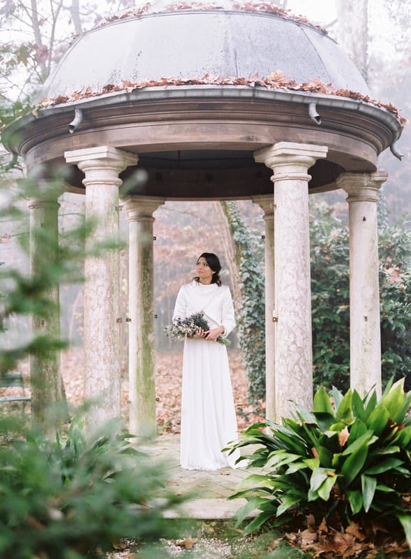 A bride stands under a gazebo holding her bouquet. She is in the middle of a beautiful garden and the sky is overcast. She wears a long-sleeved white gown and has dark hair.