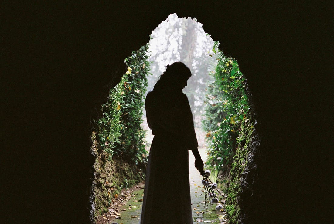A bride is walking into the garden in silhouette. She is holding a small bunch of flowers.