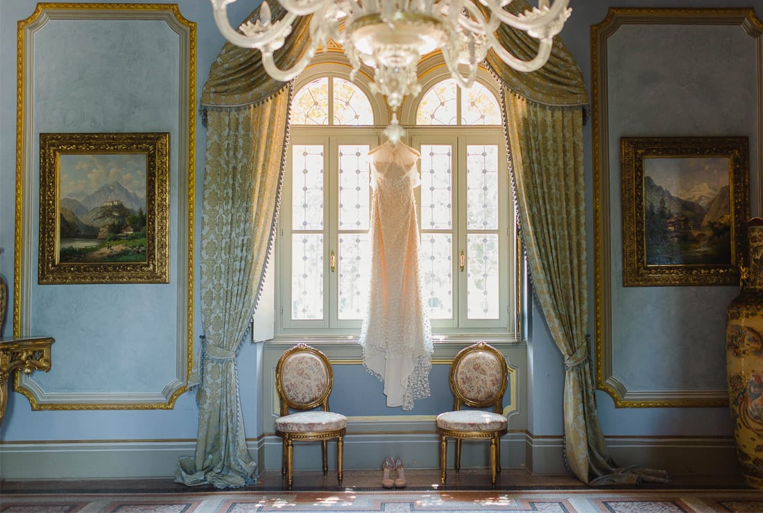 The brides gown hangs in the natural light of two window. The walls are a blue color and there is a while chandelier in the foreground.