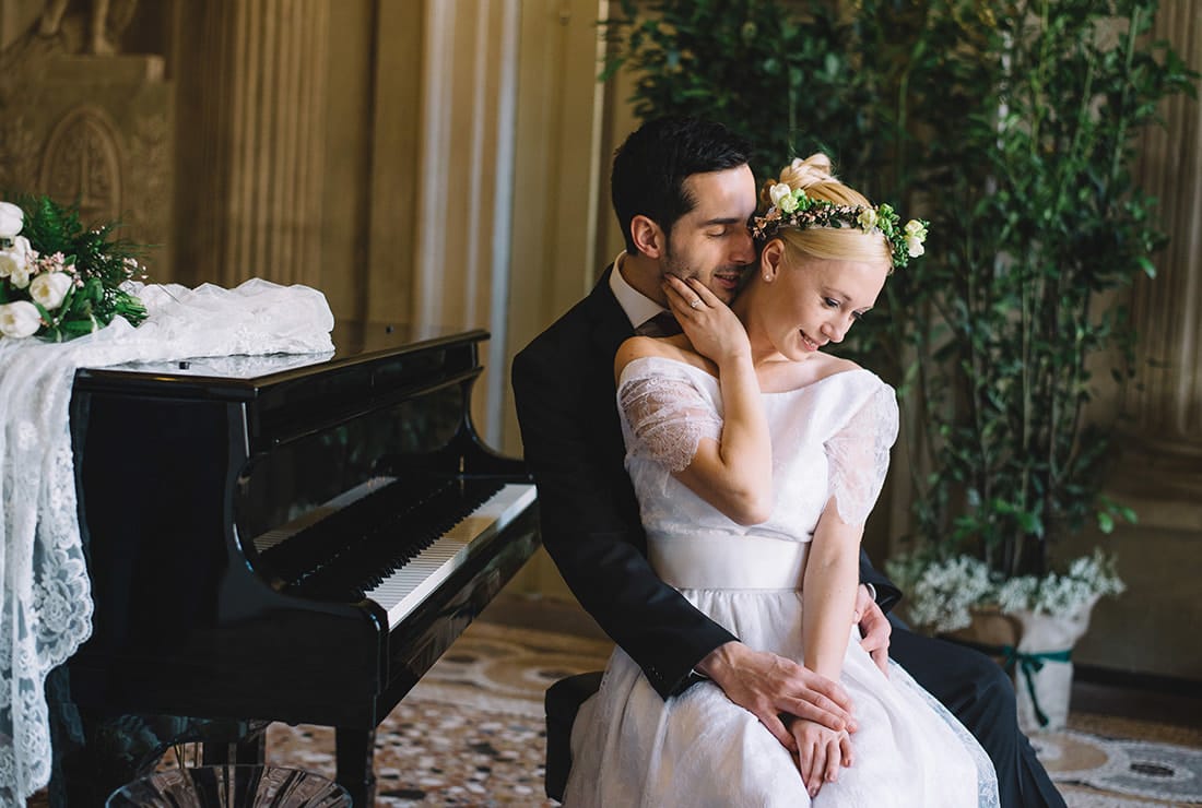 Bride and groom sit together on a piano bench in front of a grand piano. There is a lace table clothe on the top of the piano with the brides bouquet sitting in the middle.
