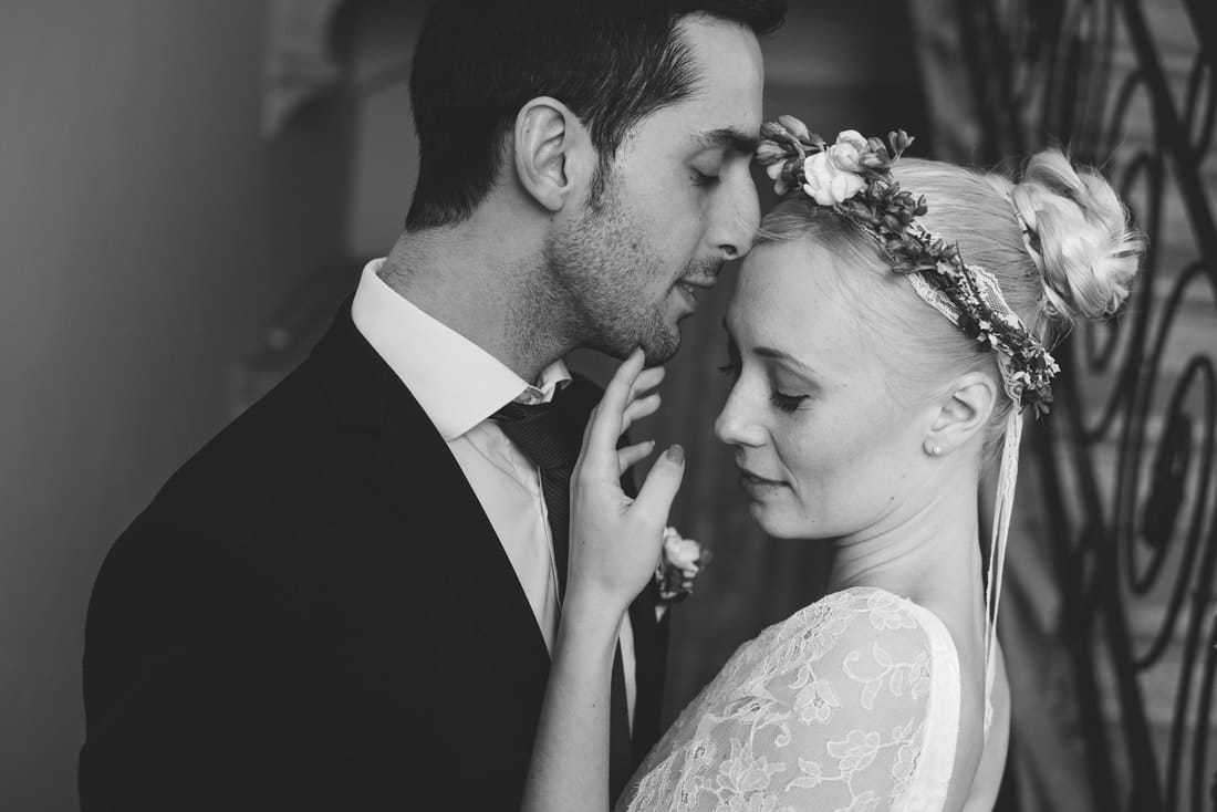 Bride and groom in an embrace. The groom is looking at the bride while she is looking away. She wears a flower crown in her hair and he is wearing a dark suit with white shirt.