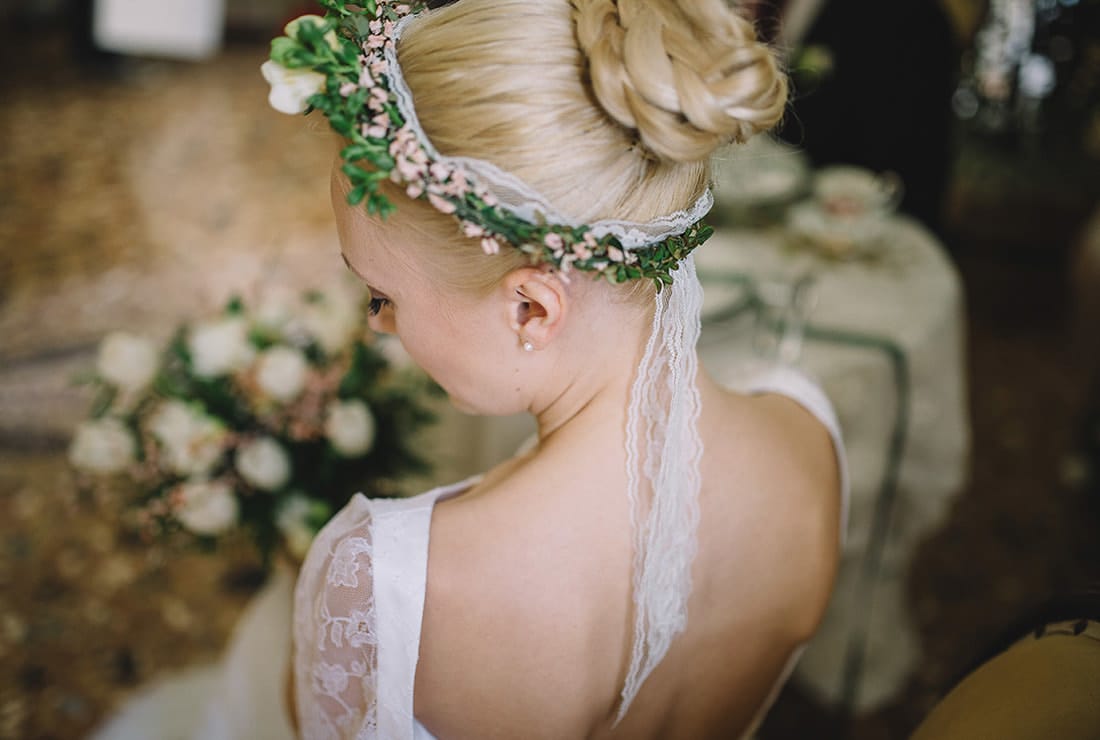 blonde bride with a braided bun in her hair wears a flower crown with lace. She is looking down as she holds her bridal bouquet.