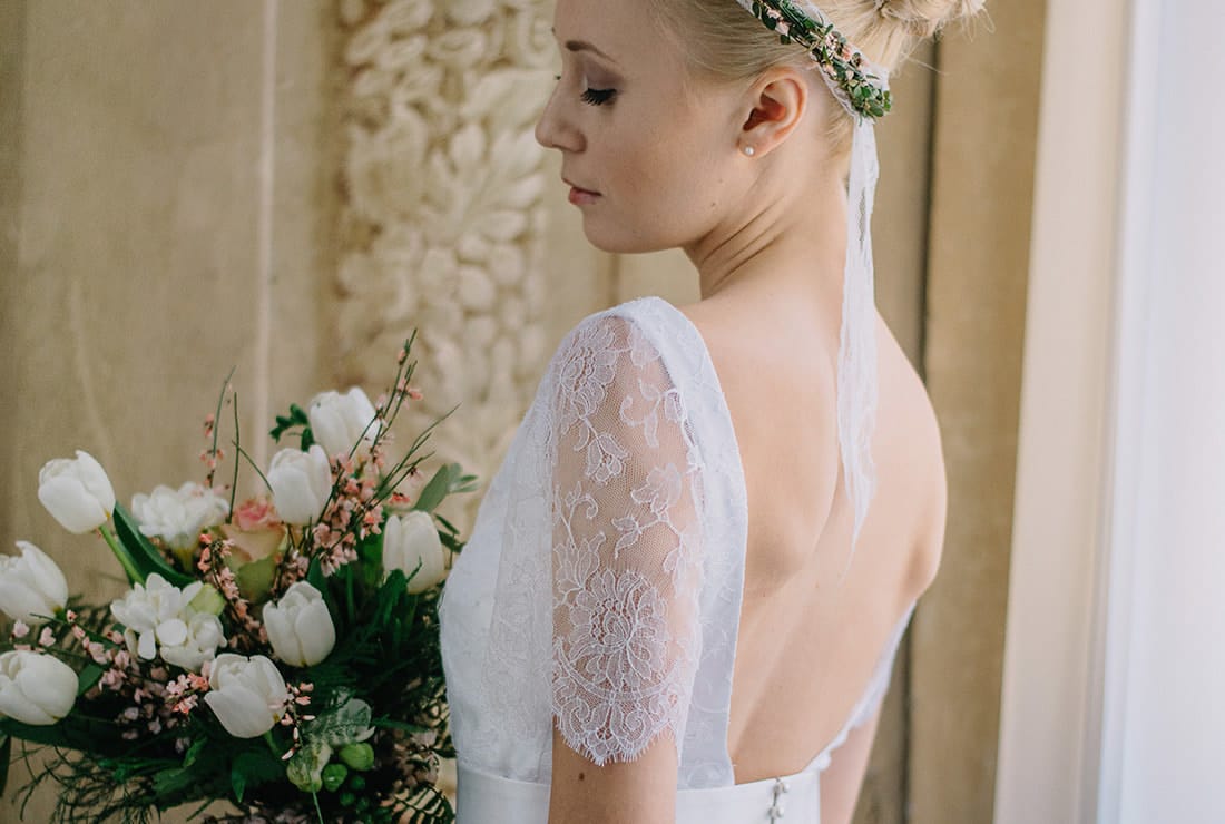 A bride looking down at her tulip and greenery bridal bouquet. She is wearing a gown with lace sleeves and a flower crown in her hair.
