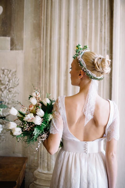 A picture of a bride looking away. Her dress has a low, open back and she is wearing a flower crown in her hair. She holds her tulip bouquet to one side.