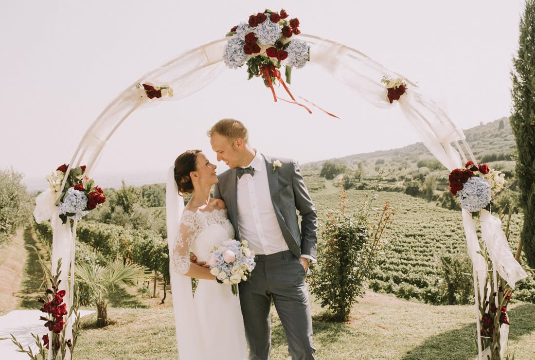Bride and groom under their ceremony arch. Red flowers adorn it with the Italian vineyard, lush and green, in the background.